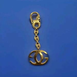 May include: A gold-toned metal keychain featuring a lobster clasp, a short chain link, and a circular pendant with an openwork design of two interlocking G letters. The word GUCCI is engraved on the clasp mechanism of this metallic accessory.