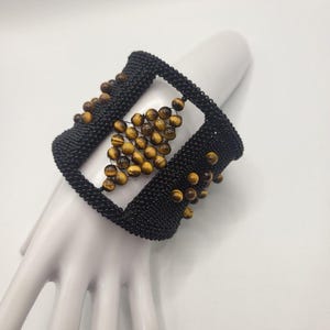 May include: A wide cuff bracelet featuring a black woven mesh structure with a central open rectangular cutout. The design is accented with round, golden-brown tiger eye beads arranged in a diamond pattern in the center and along the edges.