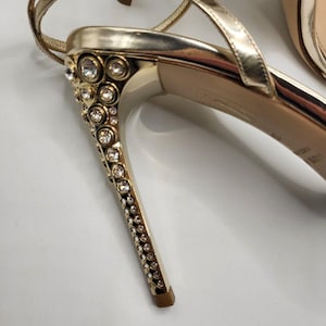 May include: A metallic gold high-heeled sandal featuring a stiletto heel embellished with numerous clear, round crystals. The shoe includes thin gold ankle straps and a smooth, light-colored inner sole with visible branding text near the heel.