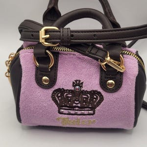 May include: A small lavender terry cloth handbag with dark brown faux leather trim, gold-tone hardware, and a dark brown adjustable strap. The front features an embroidered dark brown crown logo with a small crystal accent and the text Juicy.