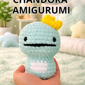 Pode incluir: Figura amigurumi de crochê azul claro com uma coroa amarela e um desenho em forma de coração branco. A figura tem olhos pretos e boca preta. O texto "CHANDORA AMIGURUMI" está no topo da imagem.