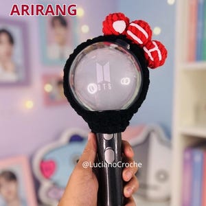 May include: A black ARMYBOMB cover with red and white crochet accents. The cover is designed to fit a BTS light stick, with the BTS logo visible. The text "ARMYBOMB COVER BTS ARIRANG" is displayed at the top. The handle is held by a hand.