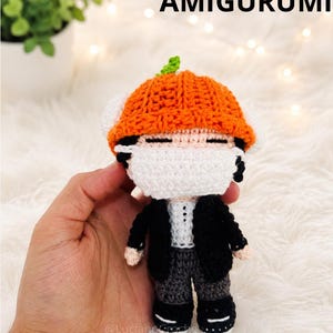 Può includere: Bambola amigurumi fatta a mano con cappello arancione e giacca nera. La bambola indossa una mascherina bianca e pantaloni grigi. Il testo "YOONGI AIRPORT AMIGURUMI" è in alto e "CROCHET PATTERN" in basso.