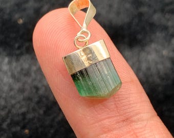May include: A pendant featuring a raw, vertically striated green tourmaline crystal capped in polished gold-tone metal. The stone transitions from a deep forest green at the base to a lighter, translucent mint green near the gold bail attachment.