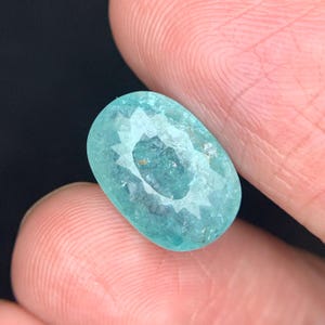 May include: A faceted, oval-shaped gemstone with a light teal color. The gem is held between two fingers, showcasing its polished surface and internal facets. The gem's cut creates a starburst effect in the center.