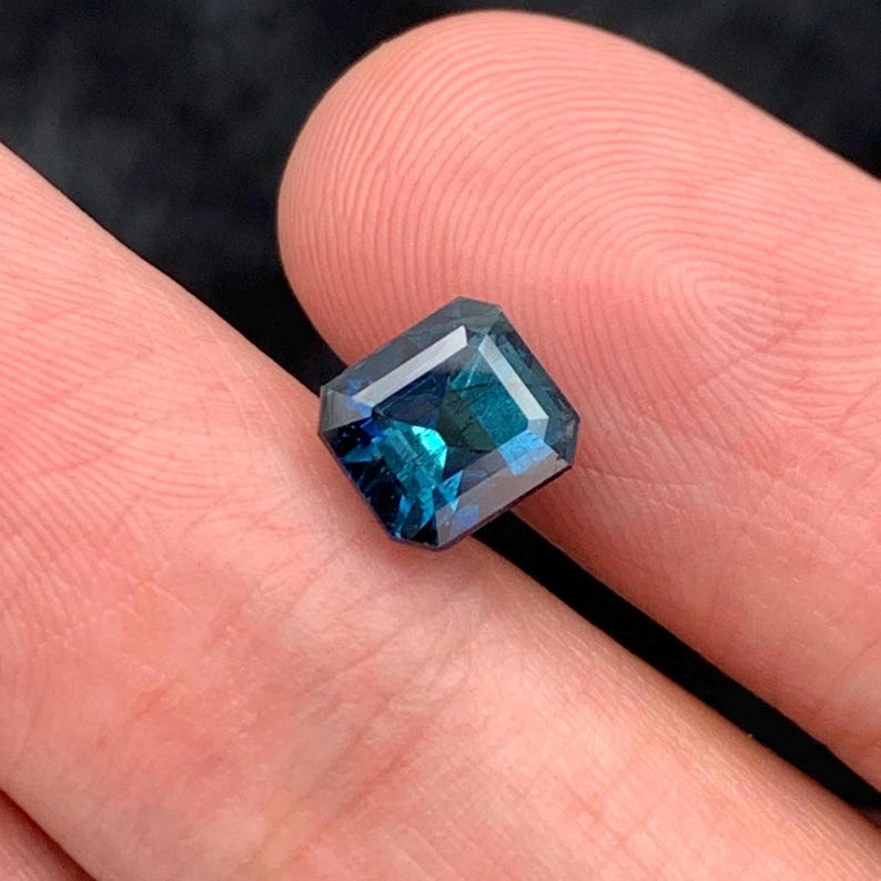 May include: A deep blue emerald cut gemstone with a slight green hue.
