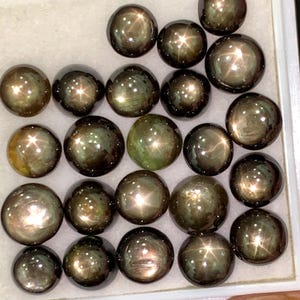 Pode incluir: Uma coleção de pedras preciosas escuras e redondas com um efeito de estrela. As gemas são uma mistura de verde escuro e castanho, com uma superfície polida e refletora. Estão dispostas numa bandeja branca, mostrando os seus padrões estelares únicos.