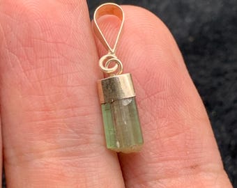 May include: A gold pendant featuring a light green tourmaline crystal. The crystal is cylindrical with vertical lines. The pendant has a loop for a chain. The crystal is approximately 1.3 cm long.