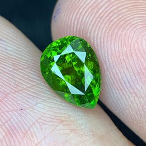 May include: A faceted, pear-shaped gemstone with a vibrant, bright green color. The gem has a complex, geometric cut that reflects light, creating a sparkling effect. The stone is held between two fingers, showcasing its size and clarity.