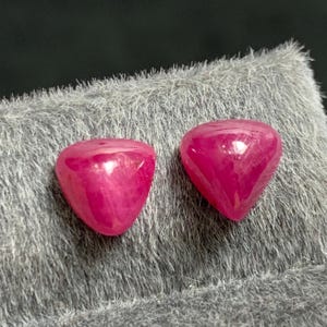 Puede incluir: Dos gemas pulidas en forma de corazón, de un vibrante tono magenta. Las gemas son lisas y reflectantes, sobre un fondo suave y gris texturizado. Las gemas parecen ser rubíes, piedras preciosas.