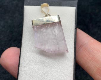 May include: A pendant featuring a light pink, rectangular crystal with a silver-colored metal cap and bail. The crystal has a natural, textured appearance. The pendant is displayed on a white surface, with a dark background.