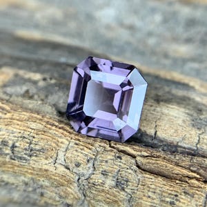 May include: A faceted, rectangular-cut gemstone with a light purple hue. The gem has a square shape with beveled edges, reflecting light. The gemstone is set against a textured, weathered wooden surface.