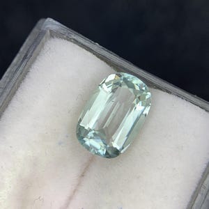 May include: A rectangular, light blue gemstone with a faceted surface. The gem is displayed in a clear plastic case against a white background. The gemstone's color is a pale, translucent blue-green.