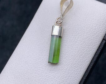May include: A silver pendant with a green tourmaline crystal. The crystal is encased in a silver setting with a loop for a chain. The pendant is displayed on a white surface.
