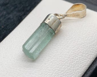 May include: A pendant with a pale green, cylindrical gemstone, silver-coloured cap and bail. The translucent gemstone is set against a white surface. The pendant is a piece of jewellery.