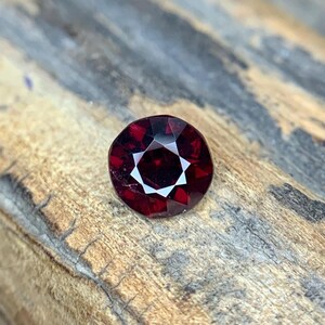 May include: A faceted, round, deep red gemstone with a star-like pattern in the center. The gem is set against a textured, wooden surface, showcasing its brilliance and color. This gemstone is ideal for jewelry making.