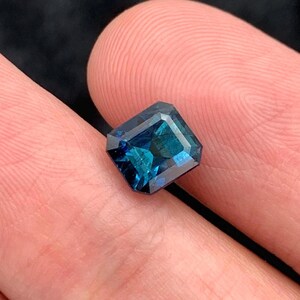 May include: A deep blue gemstone with a square emerald cut. The gemstone is set in a ring.