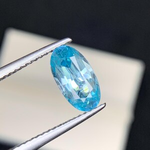 May include: A light blue, oval-cut gemstone held by tweezers. The gem has multiple facets, creating a sparkling effect. The colour is a vibrant, light blue. The background is blurred, focusing attention on the gem's clarity and colour. The gem is likely a precious stone.