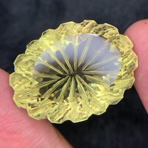 Puede incluir: Una gema facetada de forma ovalada con un tono amarillo. La gema presenta un patrón radial que emana del centro, con un borde ondulado. La gema se sostiene entre dos dedos sobre un fondo oscuro.