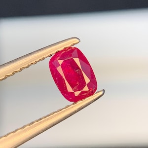 Könnte beinhalten: Ein leuchtend roter Edelstein, gehalten von einer goldenen Pinzette. Der kissenförmig geschliffene Rubin zeigt Facetten, die das Licht einfangen und seine tiefe Farbe und Klarheit zur Geltung bringen. Der Edelstein ist ein Blickfang und deutet auf Schmuck oder ein Sammlerstück hin.