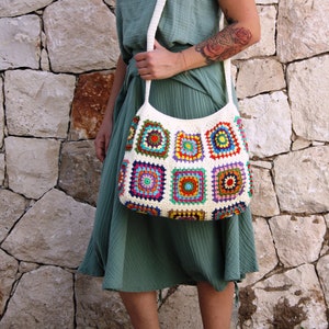 May include: A white crochet tote bag with colorful granny squares. The bag has a long strap and is worn over the shoulder.