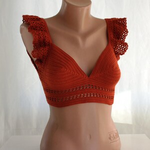 May include: A rust-colored crochet top with a V-neck and ruffled straps. The top is made with a delicate open stitch pattern.