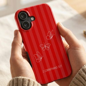 May include: A red smartphone case featuring a vertical striped pattern, three white line-art butterfly illustrations, and the text 2 CORINTHIANS 5:17 printed in white. The case has a cutout for a dual-lens camera system on the upper left corner.