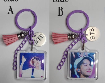 May include: A keychain with a purple ring, chain, and tassel. The keychain has a clear square pendant with a photo of a person. A silver charm with the letters "BTS" is attached.