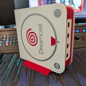 Może przedstawiać: Biało-czerwona konsola Dreamcast z logo Dreamcast z przodu. Konsola stoi na czerwonym stojaku. Posiada przycisk zasilania i kilka portów z boku. W tle widoczna jest klawiatura i monitor.