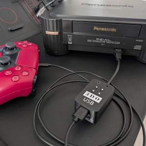 May include: A black Panasonic REAL 3DO Interactive Multiplayer console with a red game controller connected via a black cable. A black 3DO USB adapter is also connected. The console is on a black surface.