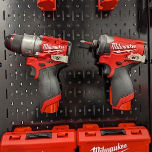 May include: Two Milwaukee FUEL cordless power tools, a drill and an impact driver, are hanging on a black pegboard. Both tools are red and black with the Milwaukee logo and FUEL branding. The tools are accompanied by their respective red toolboxes.