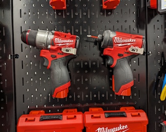 Könnte beinhalten: Zwei Milwaukee FUEL Akku-Elektrowerkzeuge, eine Bohrmaschine und ein Schlagschrauber, hängen an einer schwarzen Lochwand. Beide Werkzeuge sind rot und schwarz mit dem Milwaukee-Logo und dem FUEL-Branding. Die Werkzeuge werden von ihren jeweiligen roten Werkzeugkästen begleitet.