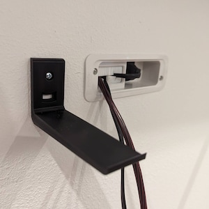 Puede incluir: Un soporte de estante de metal negro montado en una pared blanca con dos cables negros que salen de una caja de toma de corriente blanca.