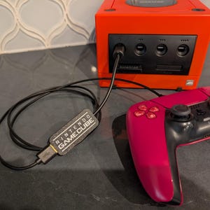 Może przedstawiać: Czerwona konsola Nintendo GameCube z podłączonym czarnym kontrolerem. Konsola ma czarny panel przedni ze szczeliną na płytę z grą i szczeliną na kartę pamięci. Kontroler to różowo-czarny bezprzewodowy kontroler z czarnym kablem łączącym go z konsolą. Kabel ma czarną etykietę z napisem "Nintendo GameCube".