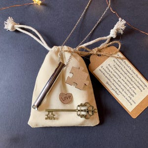 May include: A silver necklace with a small, cylindrical pendant and a key charm. The necklace is packaged in a white drawstring bag with a wooden puzzle piece and a card that reads "Love Pocket to Ease the Cravings... A necklace to support you in breaking the habit. Drawing mindful breaths. A jigsaw piece to remind you this is a journey, one piece at a time. A key to unlock your inner strength & a heart that represents self-love, self-care and the positive changes you are making. You've got this!"
