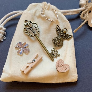 May include: A cream drawstring bag containing charms. Includes a silver ring, a bronze key, an angel figurine, a four-leaf clover, a wooden peg, and a wooden heart with "Love" engraved.