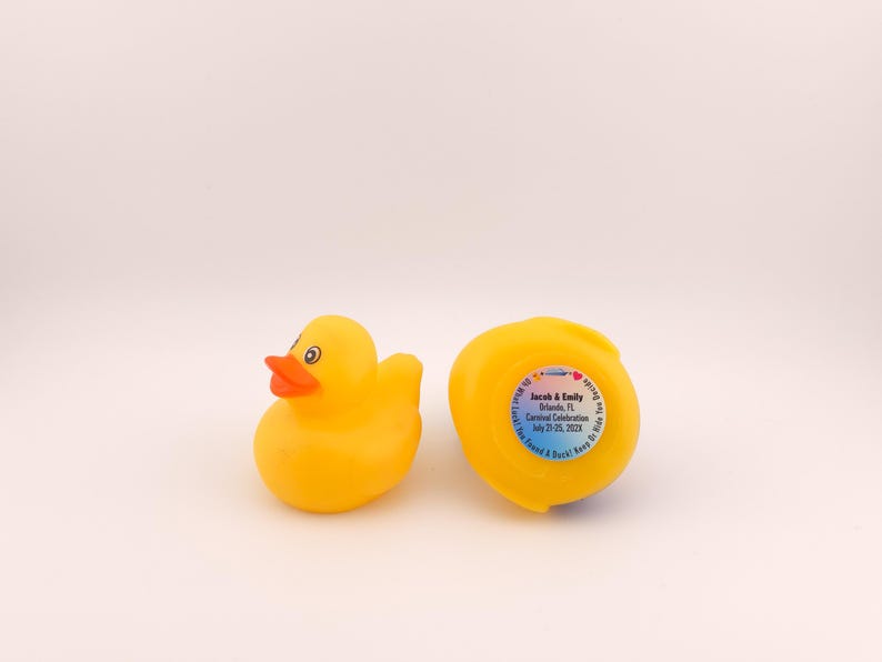 May include: Two yellow rubber duck toys. One duck is upright, with an orange beak and black eyes. The other duck is on its side, with a sticker that reads "Jacob & Emily Orlando, FL Carnival Celebration July 21-25, 2023".