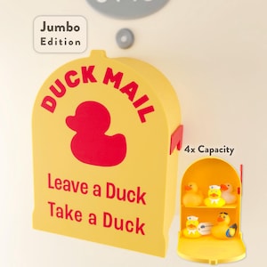 Puede incluir: Un buzón amarillo "Duck Mail" con texto rojo y una silueta de pato rojo. El texto dice "Leave a Duck, Take a Duck". Se adjunta un estante amarillo más pequeño con cuatro patos de goma. Las palabras "Jumbo Edition" están en un cuadro blanco.