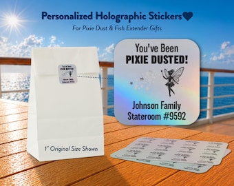 May include: A white paper bag with a square holographic sticker featuring a fairy graphic and the text "You've Been PIXIE DUSTED! Johnson Family Stateroom #9592". A sheet of identical 1-inch stickers is displayed alongside the bag on a wooden surface.