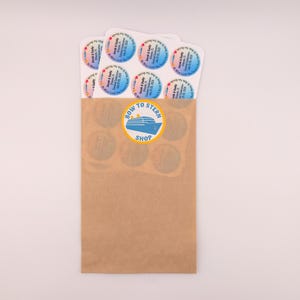 May include: A brown paper bag with a "Bow to Stern Shop" logo and several round stickers. The stickers have a colorful gradient background and text. The bag contains several stickers with the names "Sarah & Kelly" and a date.