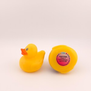 May include: Two yellow rubber duck toys. One duck is upright, with an orange beak and black eyes. The other duck is upside down, with a pink sticker that has text on it. The text on the sticker is blurry.