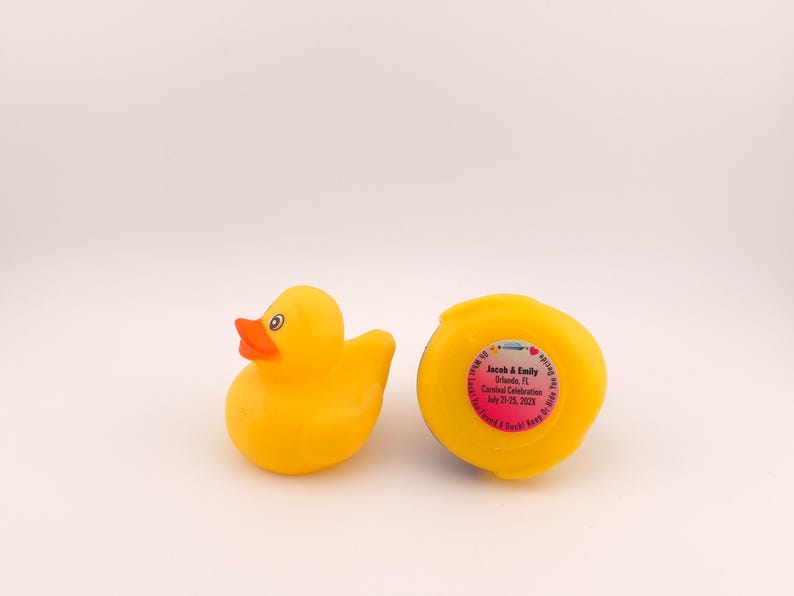 May include: Two yellow rubber duck toys. One duck is upright, with an orange beak and black eyes. The other duck is upside down, with a pink sticker that has text on it. The text on the sticker is blurry.