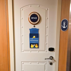 May include: A white door with a blue Duck Exchange box. The box has two rubber ducks inside. Above the box is a circular sign with the number 8143. An anchor sign is on the right.