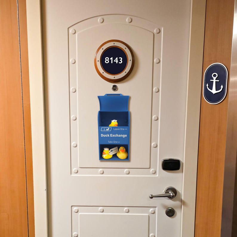 May include: A white door with a blue Duck Exchange box. The box has two rubber ducks inside. Above the box is a circular sign with the number 8143. An anchor sign is on the right.