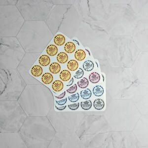 May include: Three sheets of round stickers with different designs. The top sheet has yellow stickers with red text. The middle sheet has pink stickers with black text. The bottom sheet has blue stickers with white text.