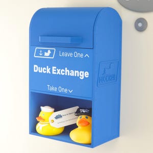 Puede incluir: Una caja azul de "Duck Exchange" con las palabras "Duck Exchange" y "Take One" impresas en la parte delantera. Dos patos de goma amarillos están dentro de la caja. Un pato lleva un sombrero de marinero. La caja está montada en una pared blanca.