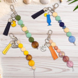Könnte beinhalten: Schlüsselanhänger mit bunten Perlen, Quasten und Anhängern. Die Schlüsselanhänger weisen eine Vielzahl von Farben auf, darunter Gelb, Grün, Blau und Orange. Zu den Anhängern gehören eine Gummiente, ein Cocktailglas, ein Boot und ein Anker.