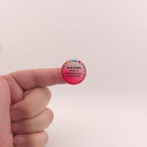 May include: A round sticker with a pink and blue gradient. The sticker has text that reads "Jacob & Emily Orlando, FL Carnival Celebration July 21-25, 202X". The sticker is held by a finger.