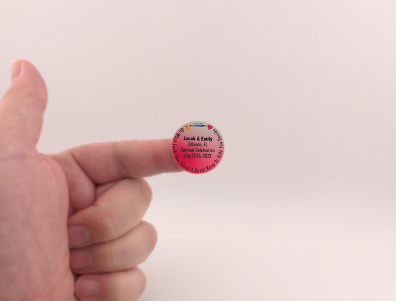 May include: A round sticker with a pink and blue gradient. The sticker has text that reads "Jacob & Emily Orlando, FL Carnival Celebration July 21-25, 202X". The sticker is held by a finger.
