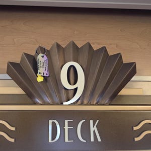 Puede incluir: Un letrero de metal marr&oacute;n con un dise&ntilde;o de abanico decorativo y el n&uacute;mero 9 en blanco. El letrero tambi&eacute;n dice "Deck" en letras blancas.